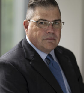 Headshot of Tim Coetzee, a stocky white man with short greying hair, glasses and a business suit and tie.